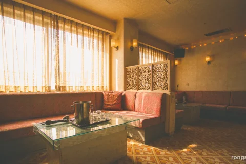 Illustration for a Chinese true crime story: the sunlit interior of an empty private karaoke room in Kunming, a long faded vermilion banquette, a low glass table with an ice bucket and a microphone, warm string lights and thin curtains, with no people